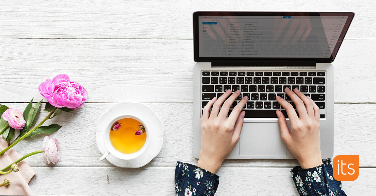 Das Bild ist von oben aufgenommen und zeigt einen Schreibtisch mit einem Laptop, einer Tasse Tee und einer rosa Blume. Die Hände einer Frau sind auf der Tastatur des Laptops platziert